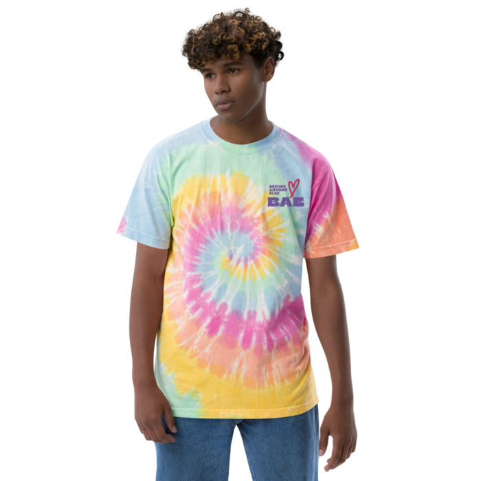 Man with curly hair, wearing a tie-dye in cotton candy colours. He is looking off to the side. he is wearing denim and there are words embroidered on the shirt that says "BAE (before anyone else)