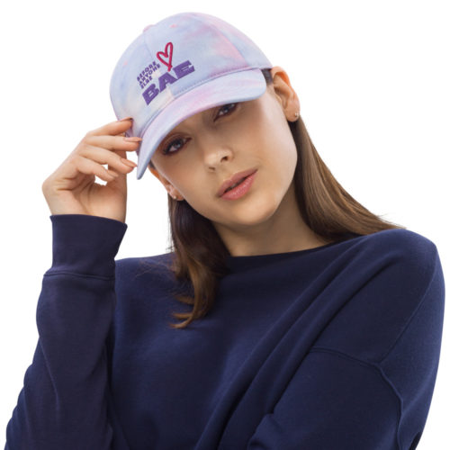 A woman with long brown hair wearing a tie-dye cotton candy coloured hat (soft pink and blue) with the words "BAE - Before Anyone Else". She is tipping her head to the side and has her hands on the hat. She is wearing a blue sweatshirt.